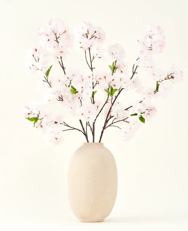Der Seiden-Kunstblumenstrauß Cherry Blossom Blush 119 cm zeigt zarte, hellrosa Kirschblüten mit grünen Blättern in einer beigen Vase, ideal als künstliche Blumendekoration vor hellem Hintergrund.